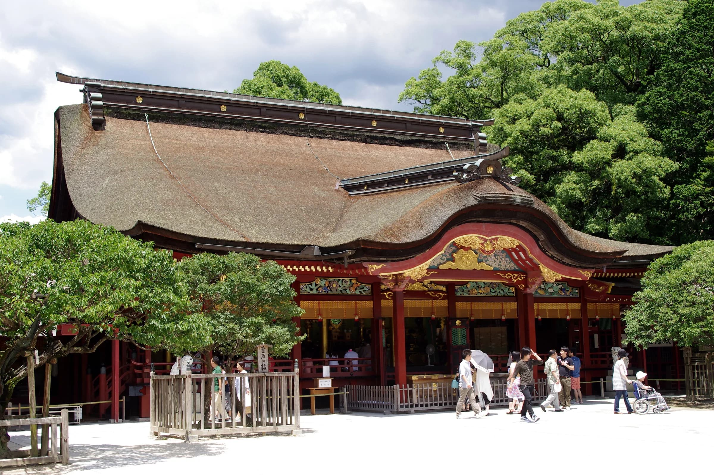 Dazaifu Tenmangu's main hall — a sweeping thatched hipped roof over red-lacquered pillars and gilded carvings, with visitors in the forecourt
