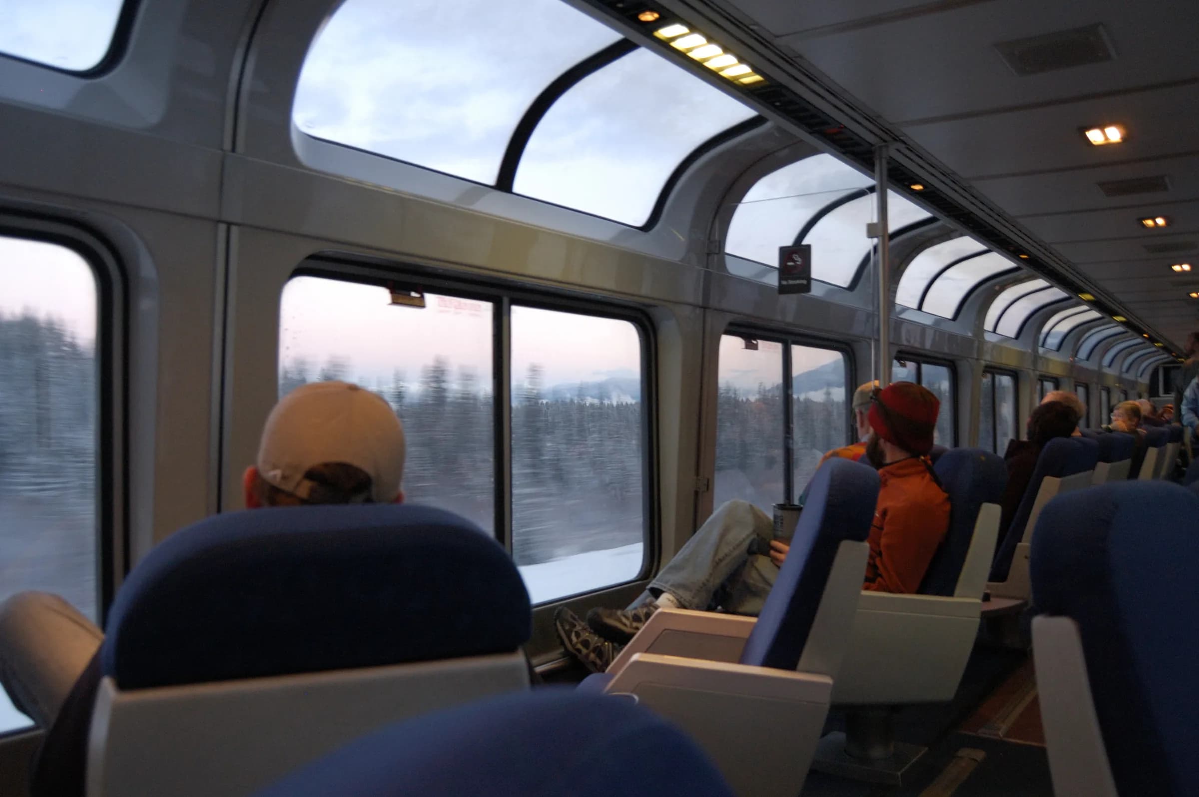 Inside an Amtrak Superliner sightseer lounge — passengers in swivel seats under the curved panoramic windows as the landscape passes